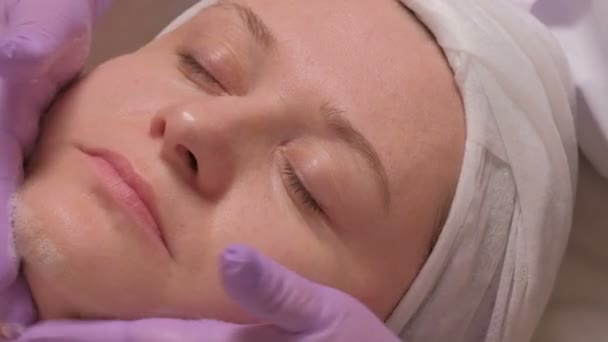 Las manos de un cosmetólogo con guantes de color lila ponen una solución de jabón en la cara femenina. Mujer de mediana edad en el procedimiento en el centro de cosmetología. Primer plano. Video crudo de alta calidad . — Vídeos de Stock