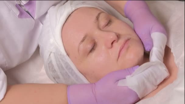 Una mujer de mediana edad está en el procedimiento de rejuvenecimiento y limpieza de la piel en la oficina de cosmetología. Las manos cosmetólogas con guantes morados limpian la cara femenina con toallitas húmedas. Vídeo crudo . — Vídeos de Stock