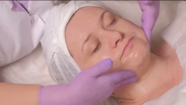 La procédure de rajeunissement et de nettoyage de la peau au bureau de cosmétologie. Gros plan. Les mains d'un cosmétologue en gants lilas mettent un gel nourrissant sur le visage d'une femme d'âge moyen . — Video