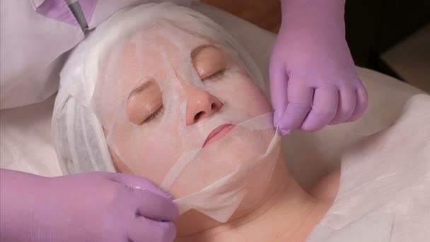Profi kozmetikus a lila kesztyű kisimítja a maszk, középkorú nő arcán. Az eljárás a bőrfiatalítás a kozmetológiai hivatalban. Spa. Közeli kép:.