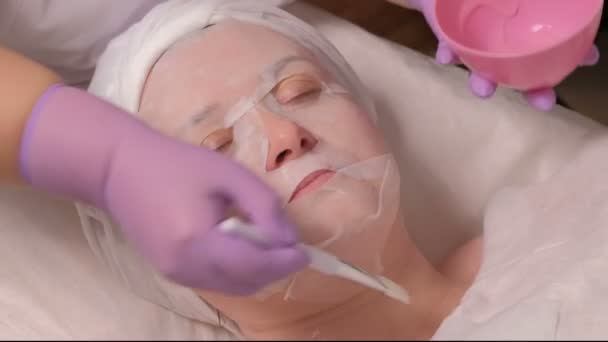 Μια γυναίκα με ένα καλλυντικό μάσκα βρίσκεται στη διαδικασία στο ιατρικό κέντρο. Ένα επαγγελματικό cosmetologist σε λιλά γάντια επιχρίσματα Θρεπτική κρέμα στο λαιμό γυναίκες μέσης ηλικίας. Αναζωογόνηση του δέρματος. Κέντρο ευεξίας και σπα. — Αρχείο Βίντεο