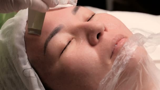 Primer plano. Mujer de mediana edad asiática se encuentra en el consultorio médico de un salón de belleza. Procedimiento cosmético para limpiar la cara con un aparato eléctrico. Rejuvenecimiento y lavado de la piel . — Vídeo de stock