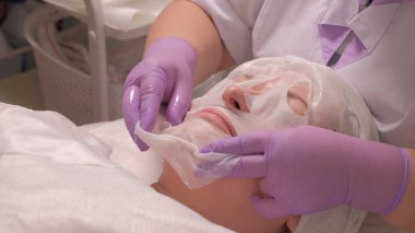 Güzellik spa ve kozmetik. Profesyonel bir güzellik uzmanı leylak eldiven orta yaşlı bir kadının yüzünde bir maske koyar. Cilt gençleştirme tıbbi ofis prosedürü.