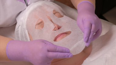 Cilt gençleştirme tıbbi ofis prosedürü. Profesyonel bir güzellik uzmanı leylak eldiven orta yaşlı bir kadının yüzünde bir maske koyar. Güzellik spa ve kozmetik.
