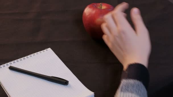 クローズ アップ 赤いリンゴは メモ帳 黒い布の上にペンの横にあります 若い男の手がテーブルから熟した果実をピックアップします — ストック動画
