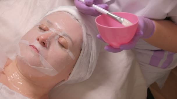 妇女在皮肤复兴做法在美容中心 美容师在白色刷子的帮助下 通过美容沙龙女性脸上的面具将凝胶涂上 医疗疗程 — 图库视频影像