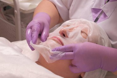 Güzellik spa ve kozmetik. Profesyonel bir güzellik uzmanı leylak eldiven orta yaşlı bir kadının yüzünde bir maske koyar. Cilt gençleştirme tıbbi ofis prosedürü.