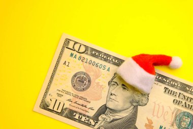 Başkan Hamilton 10 dolar üzerinde kırmızı bir Noel Baba şapkalı bize parlak sarı bir arka plan karşı bill. Noel tatil, borç, kar veya yeni yıl için indirimler maliyetine kavramı.