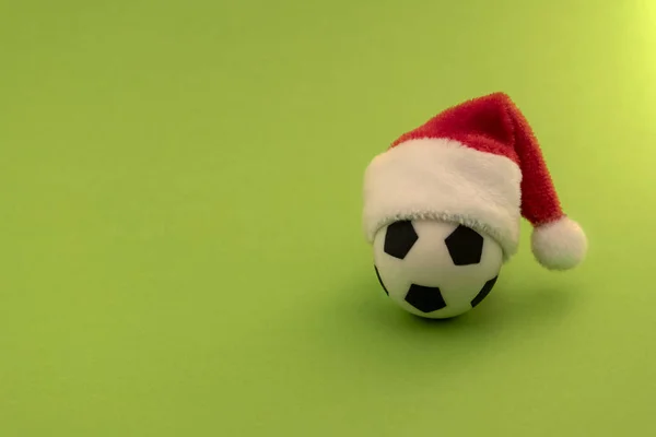 Kırmızı Noel Baba şapkalı bir futbol topu. Spor yeni yıl hediyesi kavramı. Yeşil arka plan. Kopya alanı.