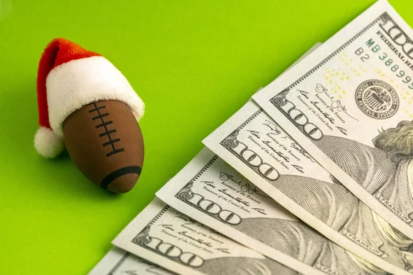 Christmas sports betting to win or lose. A red Santa Claus hat is dressed on a souvenir American football or rugby ball next to dollars on a green background. New Year\'s concept.