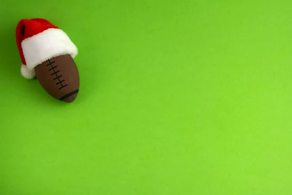 A souvenir ball for rugby or american football in a red santa claus hat on a green background. Preparation for design of tickets, posters, postcards or business cards on the subject of the sports championship. Copy space.