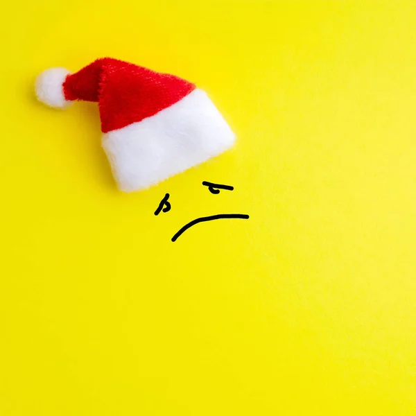 Emotion of sad face expression and red santa claus hat on yellow background. Concept of a miserable new year and christmas. Square drawing. Copy space.