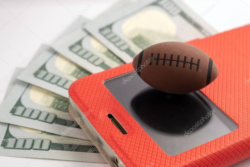 Five hundred dollars usa background. The ball for rugby or American football lies on the screen of the smartphone in a red case. Bookmaker theme on mobile. Concept on sports betting.