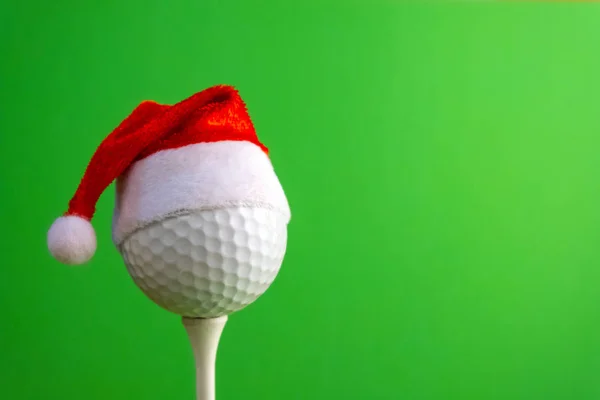 Golf ball on a tee with a red santa claus hat. Template for design greeting card for golfer for new year or christmas. Green background. Copy space.