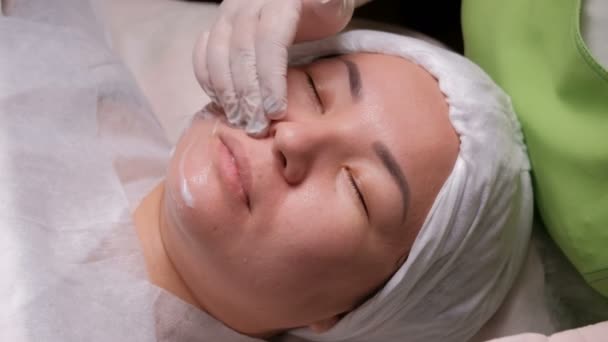 Professionele schoonheidsspecialist in witte handschoenen zet een voedende crème op het gezicht van een Aziatische vrouw. Cosmetische procedure in de schoonheidssalon. Reinigings- en verjonging van de huid in de cosmetologie Center. — Stockvideo