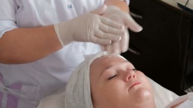 Profesyonel bir güzellik beyaz eldiven eller Close-up krem bir kadının yüzüne koyar. Kozmetoloji merkezi cilt temizliği usulü hakkında memnun kadın.