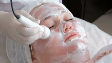 Gözleri kapalı ve bir güzellik salonu terapötik bir yordamda yüzü üzerinde krem olan kadın. Bir profesyonel güzellik uzmanı veya doktor ultrason cihazları ile kadın yüzü acupressure gerçekleştirir.
