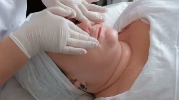 Europeiska eller amerikanska kvinna på en hudrengöring förfarande vid en kosmetologi center. Närbild av händerna på en professionell kosmetolog i vita handskar soaping ett kvinnligt ansikte. — Stockvideo