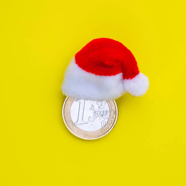 The concept of financial costs of the European New Year and Christmas. Coin one euro in a red santa claus hat on a yellow background. Square frame Copy space.