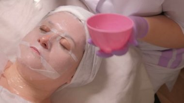 Onun gözleri kapalı yalanlar üzerine bir gençleştirici kozmetik prosedür olan orta yaşlı bir kadın. Lila eldiven bir güzellik onun elinde pembe bir fincan ile bir jel istemcinin yüz beyaz bir fırça ile uygulanır.