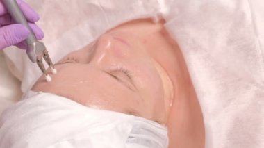 İki elektrot yardımıyla istemcinin alnına Microcurrent acupressure. Kapalı gözlerle orta yaşlı bir kadın yüzü ile şeffaf bir jel sürülür. Donanım kozmetik salonda
