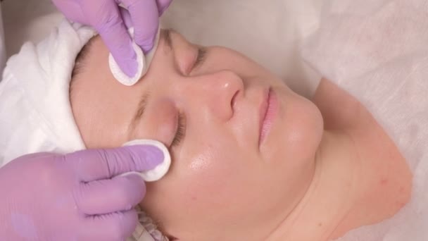 クローズ アップ 美容師は 彼女の目と中年女性の顔を閉じる綿のパッドを使用して紫手袋ワイプで手します ビューティー サロンで肌の若返りの手順 — ストック動画