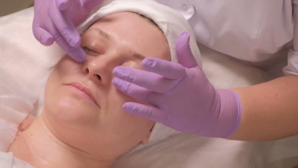 在淡紫色手套的美容师的手把奶油套在眼睛上 贴在中年妇女的脸上 美容院美容诊所的治疗程序 — 图库视频影像