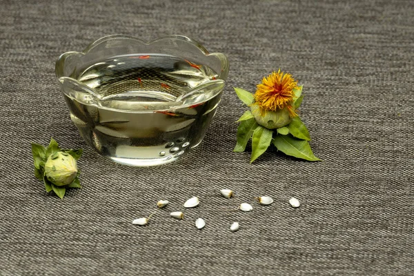Carthamus tinctorius, Amerikan safran, vahşi thistle. Aspir boyama: bud, sarı çiçek, tohum ve yağ gri kumaş bir cam fincan bir arka plan üzerinde. Carthamus tinctorius, Amerikan safran, vahşi thistle. Zamanlardan beri yağlı tohum ve boyama, kültür 
