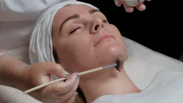 De procedure voor het reinigen van de huid van het gezicht in een schoonheidssalon of cosmetologie center. Close-up van een cosmetische penseel verspreiding crème op de wangen van een mooi meisje. Niet-afgehandelde video. — Stockvideo