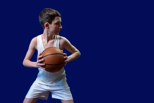 一名身穿白色 T恤和运动短裤的少年拿着篮球 把目光移开 复制空间 体育海报的空白 在蓝色背景上隔离 — 图库照片