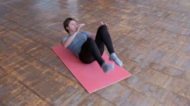 Atletik kız gri tişört ve siyah tayt pembe bir halının üzerinde yatıyor ve dönüşümlü olarak karın kasları güçlendirmek için bacaklarını görevini yerine getirmektedir. Fitness vücut esnek. Spor salonu zemin üzerinde eski parke.