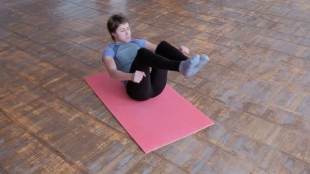 Fitness test flex. Gyakorlat, hogy a hasi izmok erősítésére. Egy nő sportoló egy szürke pólót és fekete harisnya fekszik egy rózsaszín szőnyeg, és hajlik a lábát. A tornaterem padlón régi parketta. — Stock videók