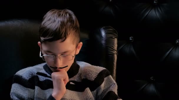 Молодой человек в полосатом свитере сидит в черном кресле. Подросток с приклеенными усами держит ручку в руке и размышляет. Крупный план . — стоковое видео
