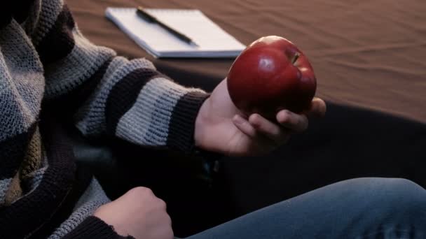 Un jeune homme en pull rayé avec une pomme rouge à la main. Un adolescent prend un bloc-notes et un stylo de la table pour enregistrer. Gros plan . — Video