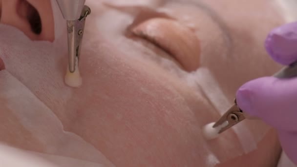 Närbild. Kosmetolog i lila handskar börjar att göra förfarandet med en föryngrande effekt med elektrisk elektroder. Kvinna med ansiktsmask i kosmetologi center. Makrofotografering. — Stockvideo
