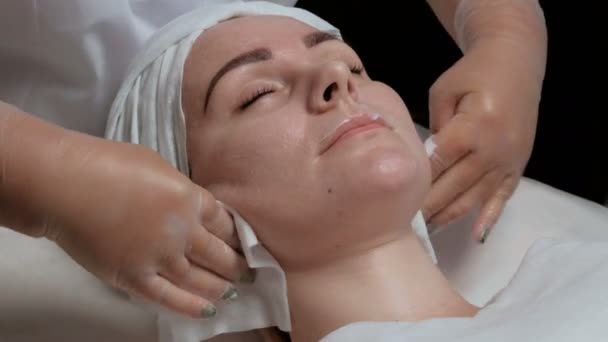Крупним планом. Білосніжний косметолог натирає шкіру пацієнтів білими серветками. Красива дівчина з кремом на обличчі в косметологічному центрі. Процедура омолодження та очищення обличчя — стокове відео