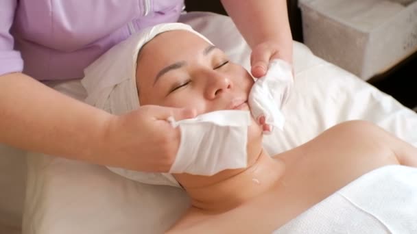 Aziatisch meisje op de procedure in de schoonheidssalon. Massage, verjonging en gezichtsreiniging. Womens handen van een professionele schoonheidsspecialist veeg de klanten huid met natte doekjes. — Stockvideo