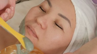 Profesyonel bir Kozmetolog, Asyalı bir kadının yüzüne bir aljinat maskesi uygular. Close. Güzellik salonunda oryantal bir kız için gençleştirme prosedürü. Renk düzeltmesi olmadan RAW video.