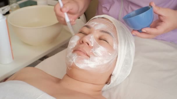 美容师在一位亚洲中年妇女的脸上涂上按摩膏 在美容院恢复活力的程序 女性手中的白色美容刷和蓝色橡胶杯 — 图库视频影像