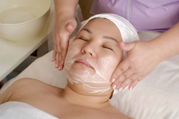 Esteticista do dia. Uma menina de aparência asiática em um salão de beleza. As mãos de um cosmetologista colocar sabão branco suds no rosto de uma mulher oriental. O procedimento de limpeza e rejuvenescimento da pele . — Fotografia de Stock