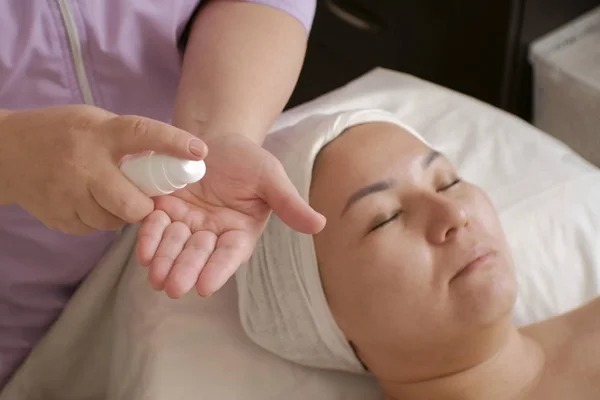 Een meisje van Aziatische verschijning in een schoonheidssalon. Een professionele schoonheidsspecialist knijpt uit een witte fles cosmetisch product op de hand. Cosmetische procedure voor het reinigen en verjonging van de huid van de vrouw — Stockfoto