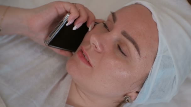 Ragazza carina con un cappello cosmetico sulla testa che parla su un telefono cellulare. Una donna è distesa in un ufficio estetiste su un letto in un salone di bellezza . — Video Stock