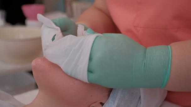 De schoonheidsspecialiste in groene handschoenen en een roze Kamerjas wrijft het gezicht van een mooi meisje met witte natte doekjes. Close-up. Cosmetische procedure voor het reinigen van de vrouwelijke huid in een schoonheidssalon. — Stockvideo