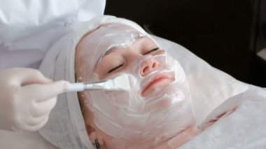 Beyaz eldiven güzellik terapisti bir fırça ile tam bir kadın yüzüne beyaz masaj kremi uygulayarak. Bir güzellik salonunda orta yaş kadın cilt sıkma gençleştirici prosedür. Close.