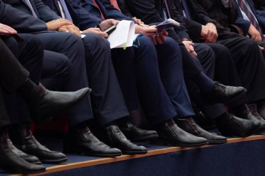 Yetkililer toplantı odasında oturuyor. Erkek pantolon ve siyah ayakkabılar bacaklar. Makaleler ve el telefonlar.