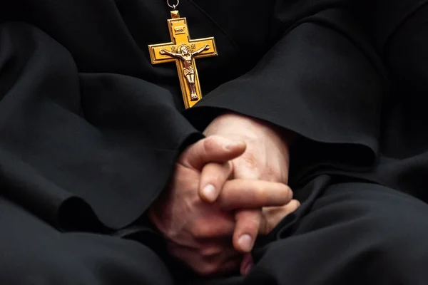 Siyah giysili bir rahip göğüs üzerinde İsa'nın çarmıha germe ile altın haçı. Ortodoks din sembolü. Eller birlikte odak dışında bulanık. Sığ derinlik-in tarla. — Stok fotoğraf