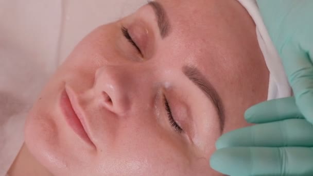 在美容院面对一个漂亮的女孩 戴蓝色手套的美容师在患者的眼睛周围涂上透明的抗皱凝胶 女性皮肤恢复和再生的程序 — 图库视频影像