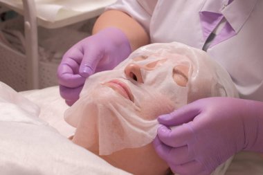 Kadın yüzünde kozmetik maske var. Leylak eldivenli profesyonel bir kozmetolojiuzmanının elleri. Gençleştirici ve temizleyici cilt prosedürü. Kaldırma ve kırışıklık karşıtı maske.