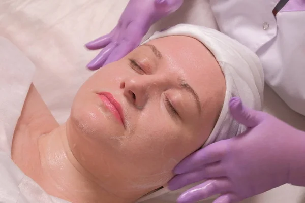 Mujer con los ojos cerrados en un procedimiento cosmético en un salón de belleza. Las manos de un cosmetólogo con guantes lila en las sienes de la cara de una mujer. Efecto relajante y rejuvenecedor de la piel . — Foto de Stock