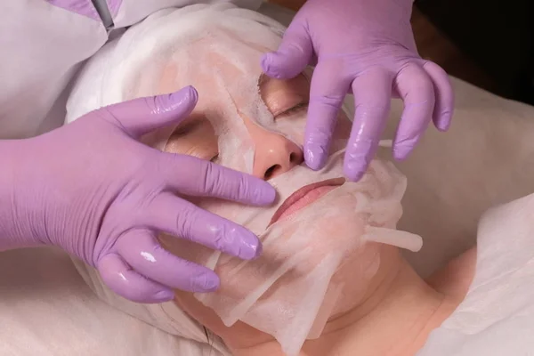 De handen van een professionele cosmetoloog in Lila Handschoenen leggen een voedend masker op de patiënt. Cosmetische masker op het gezicht van een vrouw. Huid verjongende reinigingsprocedure. Heffen en anti-rimpel — Stockfoto